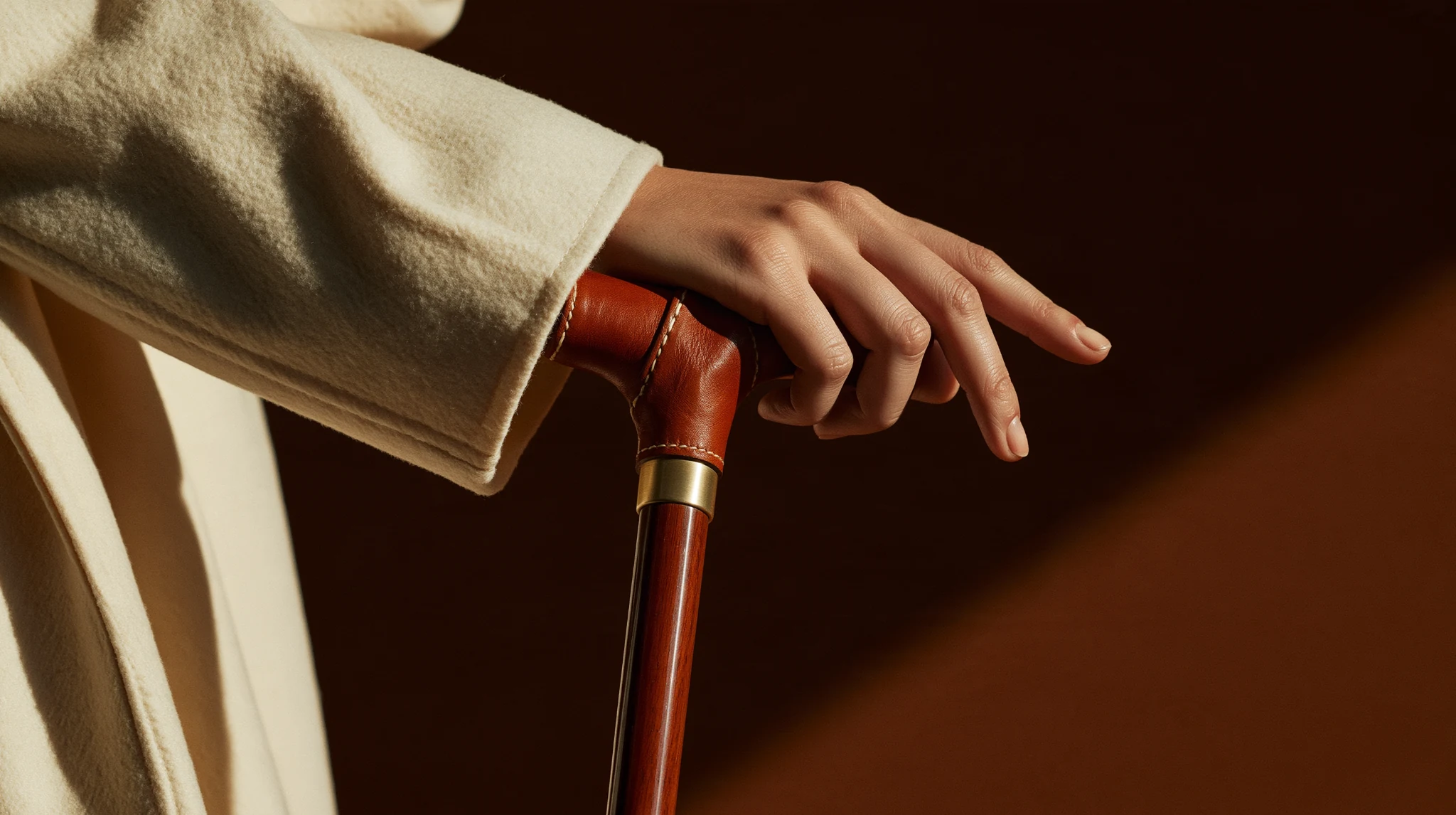 Close-up of a hand resting on a leather-handled wooden walking stick with brass detailing, beside a cream coat.